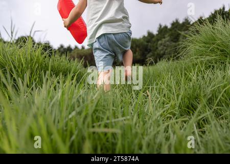 Felice concetto di infanzia, esplorando il mondo intorno a te. Adorabile ragazza che corre a piedi nudi sull'erba in un parco durante il giorno e regge una mongolfiera. Foto Stock