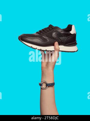 Una mano femminile con un bracciale che regge una sneaker nera alla moda con suola e bancone bianchi, isolata su uno sfondo blu. Scarpe sportive eleganti Foto Stock