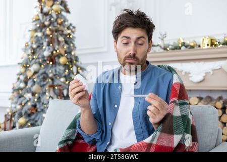 Un giovane stanco si siede malato a casa durante le vacanze di Capodanno. Seduto sul divano coperto da una coperta e guardando il termometro con sorpresa, ha un aspetto e si sente male. Foto Stock
