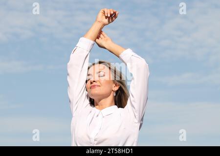 ESPRESSIONI DI FELICITÀ E BENESSERE DA UNA RAGAZZA BIONDA IN CAMICIA BIANCA CON CIELO BLU SULLO SFONDO Foto Stock