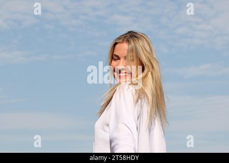 ESPRESSIONI DI FELICITÀ E BENESSERE DA UNA RAGAZZA BIONDA IN CAMICIA BIANCA CON CIELO BLU SULLO SFONDO Foto Stock