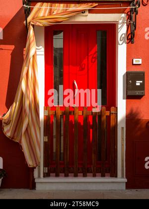 Una porta rossa brilla di luce su questa facciata di una casa a Burano a Venezia, Italia. La tenda che adorna la porta gli dà movimento. Foto Stock