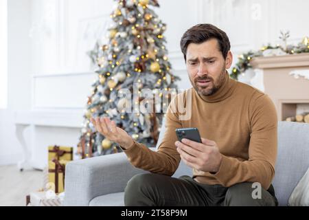 Un uomo infelice seduto vicino all'albero di Natale il giorno d'inverno, ha ricevuto cattive notizie al telefono, festeggiando il Capodanno e il Natale, lettura infelice, utilizzo dell'app sullo smartphone, soggiorno a casa sul divano. Foto Stock