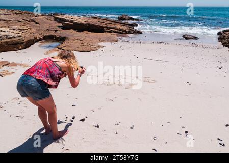 Una donna in pantaloncini denim scatta una foto di scrittura sulla sabbia a Lamberts Bay in Sudafrica Foto Stock