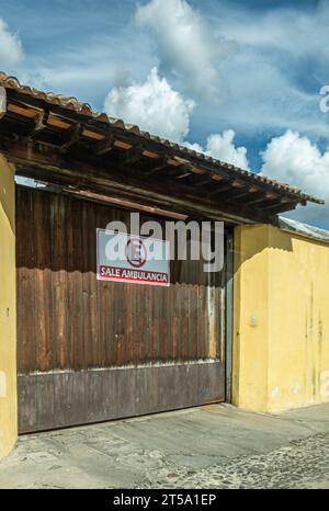Guatemala, la Antigua - 20 luglio 2023: Il segnale di uscita dell'ambulanza sul cancello marrone proibisce il parcheggio davanti. Incastonata in parete gialla sotto il paesaggio blu, S Foto Stock