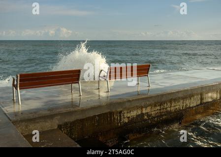 Le onde scorrono sul lungomare con due panchine. Foto Stock