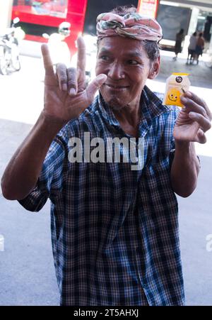 L'uomo più anziano per strada a Bangkok, Thailandia, fa un segno del gesto della mano delle corna, saluto standard del rock 'n' roll, gesto della mano dei fan del rock, corna del diavolo o corna di metallo, segno della mano "rock on". D'altra parte, ha in mano un campione gratuito di gelatina al mango appiccicosa al riso Sunsu, che un chiosco vicino sta regalando come promozione Foto Stock