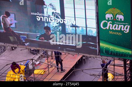 Vista dall'alto. I giovani assemblano le strutture delle bancarelle del mercato notturno di Patpong, sulla Patpong Road 1, nel distretto di Silom. Bangkok, Thailandia. Le bancarelle vengono assemblate alle 15:00 e portate fuori all'una di notte ogni giorno. Un cartello illuminato sopra i lavoratori, un cartello permanente per pubblicizzare le bevande a marchio Chang accanto a quello. Foto Stock