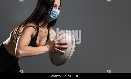 Vista laterale atletica giocatore di rugby femminile che tiene la palla mentre indossa la maschera medica Foto Stock