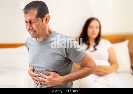 Un uomo che soffre di crampi allo stomaco seduto sul suo letto vicino a sua moglie Foto Stock