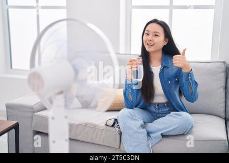 Giovane donna asiatica che beve un bicchiere d'acqua che si gode l'aria dal ventilatore sorridente felice e positiva, pollice su facendo eccellente e segno di approvazione Foto Stock