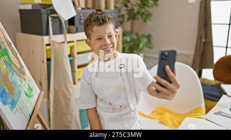 Adorabile artista biondo sorridente mentre fa un selfie con uno smartphone in un vivace studio d'arte, pennello in mano durante le lezioni di disegno. Foto Stock