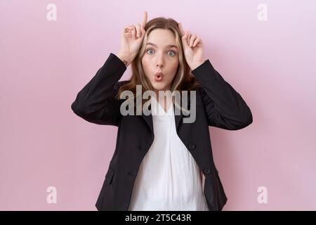 Giovane donna d'affari caucasica che indossa una giacca nera che fa gesto divertente con le dita sopra la testa come corna toro Foto Stock