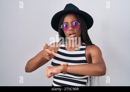 Giovane afro-americano con trecce che indossano cappello e occhiali da sole in fretta indicanti tempo di visualizzazione, impazienza, turbato e arrabbiato per il ritardo della scadenza Foto Stock