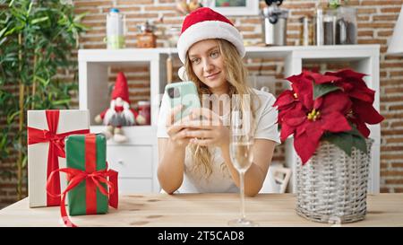 Giovane bionda che festeggia il natale con uno smartphone e beve champagne in sala da pranzo Foto Stock