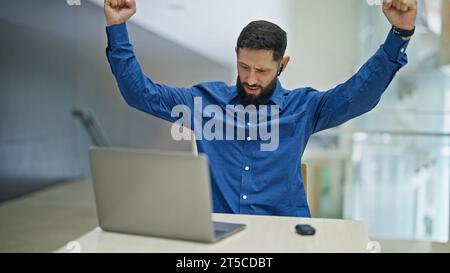 Giovane ispanico lavoratore d'affari che usa un computer portatile e auricolari per festeggiare in ufficio Foto Stock