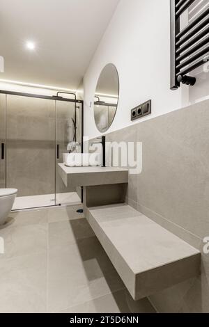Bagno bellissimo e moderno con piastrelle grigie, ripiano dello stesso materiale, specchio circolare, cabina doccia con schermi in vetro, asciugamano a parete radia Foto Stock