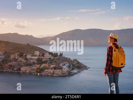 Bella donna asiatica felice con zaino che si gode le vacanze alle isole Symi al tramonto. Vista del porto di Symi o Simi, è una piccola isola del Dodecaneso, Grecia, atmosfera tranquilla e architettura favolosa. Foto Stock