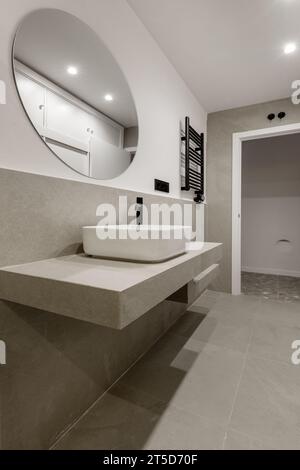 Bagno bellissimo e moderno con piastrelle grigie, ripiano dello stesso materiale, specchio circolare Foto Stock