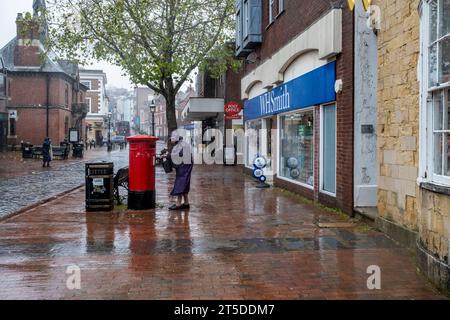 Una signora anziana pubblica Una lettera in Una casella postale durante Una doccia a pioggia, High Street, Lewes, Regno Unito Foto Stock