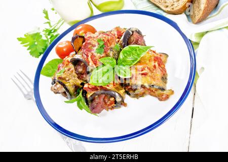 Panini di melanzane cotti al forno con carne, pomodori e formaggio in un piatto, tovagliolo, basilico, pane e forchetta su uno sfondo di legno chiaro Foto Stock