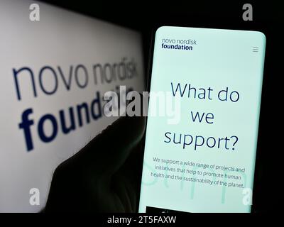 Persona in possesso di cellulare con pagina Web dell'organizzazione danese Novo Nordisk Foundation davanti al logo. Concentrarsi sul centro del display del telefono. Foto Stock