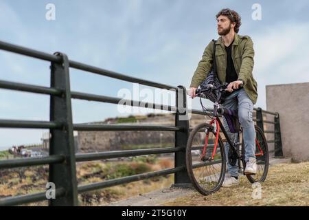 Un giovane hipster barbuto che va in bicicletta, Un giovane uomo contemplativo che pedala attraverso sentieri urbani, godendosi una piacevole corsa Foto Stock