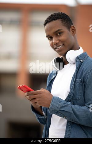 Un giovane ragazzo sorridente che guarda la fotocamera si rilassa usando il telefono.un uomo millenario che sorride e invia SMS indossando le cuffie. Foto Stock