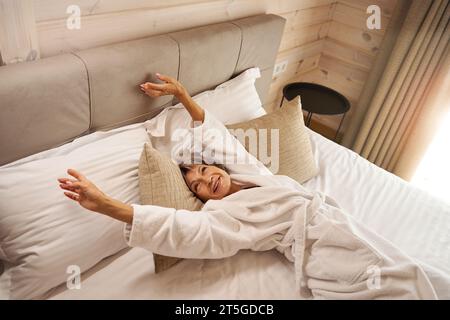 Una donna anziana e felice si sveglia in una camera da letto accogliente con morbidi cuscini Foto Stock