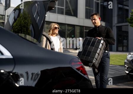 Donna d'affari aspetta mentre l'autista porta una valigia nel bagagliaio dell'auto Foto Stock