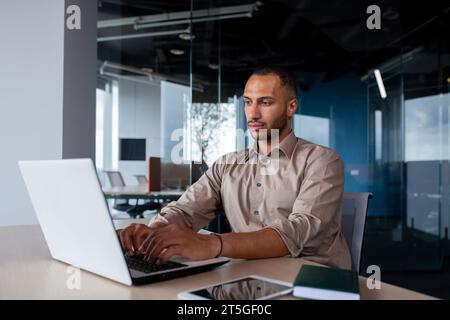 Uomo serio e concentrato che lavora all'interno dell'ufficio sul posto di lavoro, uomo d'affari che digita sulla tastiera del notebook, programmatore che sviluppa nuovo software. Foto Stock