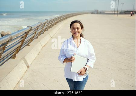 Gara mista di successo, bella donna imprenditrice freelance cammina lungo la passeggiata sull'oceano Atlantico con un notebook in mano, sorride con fiducia Foto Stock