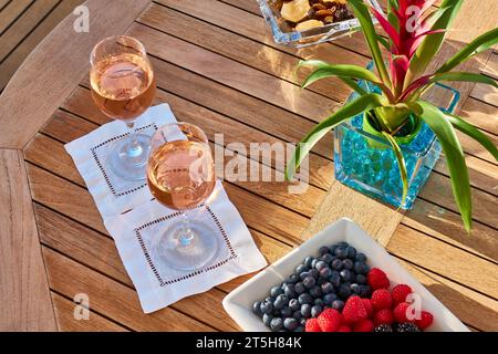 Due bicchieri di vino rosa su un tavolo rotondo in teak con un cactus fiorito e una ciotola di mirtilli, lamponi e fragole. Foto Stock