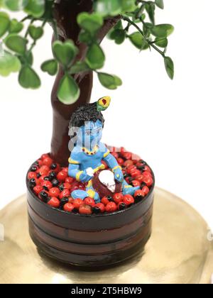 una colorata statua della bambola sri krishna in blu che mangia burro da una pentola sotto un albero, un avatar del dio vishnu della religione indù Foto Stock