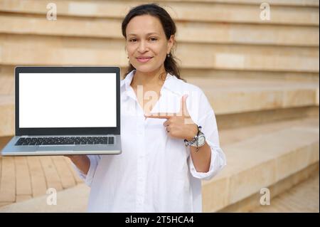 Bellissima giovane donna multietnica in camicia bianca casual, con indice puntato verso uno schermo digitale mockup bianco con spazio pubblicitario, sorridente Foto Stock