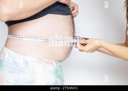 Una donna misura la pancia di un uomo in sovrappeso con un metro a nastro su uno sfondo leggero, obesità e dieta, salute Foto Stock