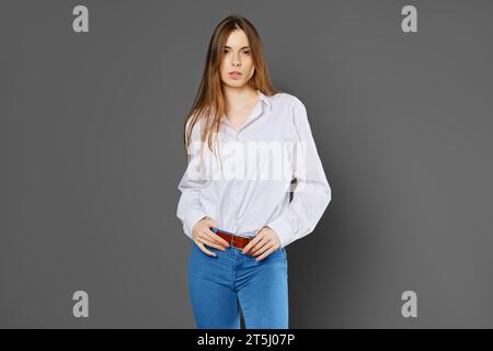 Ritratto in studio di una giovane donna in camicia bianca, jeans blu su sfondo grigio Foto Stock