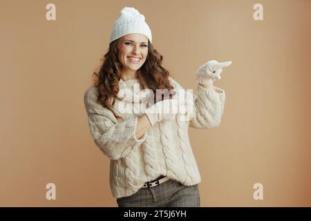 Ciao inverno. elegante donna sorridente di 40 anni con maglione beige, guanti e cappello che puntano a qualcosa su sfondo beige. Foto Stock