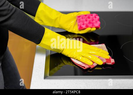 una donna con i guanti pulisce il piano cottura. lavare il piano cottura in cucina. una mano in un guanto giallo strofina il piano cottura con uno straccio. Foto Stock