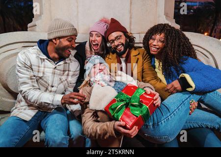 Giovane donna adulta eccitata che tiene tra le braccia un regalo di compleanno o un regalo di natale dei suoi migliori amici che ridono insieme con complicità. Felice gruppo di persone multirazziali che festeggiano la notte di Natale. Foto di alta qualità Foto Stock