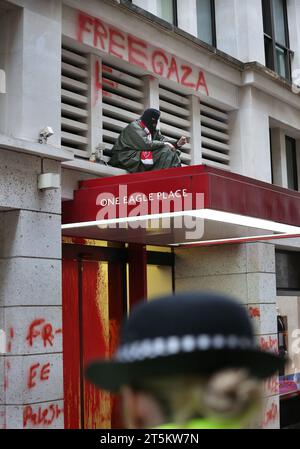 Un attivista protesta contro il baldacchino sopra l'ingresso principale degli uffici, mentre gli agenti di polizia tengono d'occhio la parte sottostante. Gli attivisti di Palestine Action occupano e prendono di mira il gigante italiano dell'industria degli armamenti Leonardo nel loro quartier generale di Londra e li chiudono. Leonardo rifornisce Israele con jet da combattimento e armi attualmente in uso a Gaza. Palestine Action chiede che le compagnie di armamento che forniscono armi a Israele chiudano definitivamente. Hanno annunciato che le società che vendono armi alla forza di difesa israeliana e alle loro società partner saranno prese di mira con azioni dirette. Queste azioni ar Foto Stock