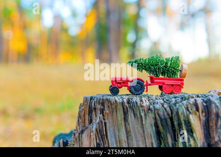 Il trattore giocattolo trasporta un piccolo albero di Natale arroccato in cima a un vecchio ceppo di alberi nel cuore della foresta. Concetto di registrazione industriale. Foto Stock