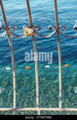 Lucchetto a forma di cuore su una vecchia recinzione metallica da un primo piano trasparente della baia marina. Forte simbolo d'amore sulla griglia contro l'acqua dell'oceano nelle giornate di sole. Artefatto carino Foto Stock