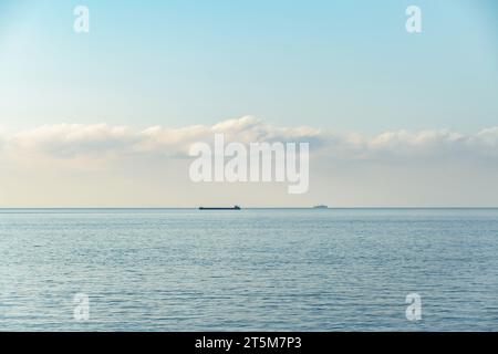 Tranquilla vista sul mare con due navi visibili all'orizzonte la mattina presto. Il mare calmo sotto il cielo blu punteggiato di nuvole crea un'atmosfera tranquilla Foto Stock