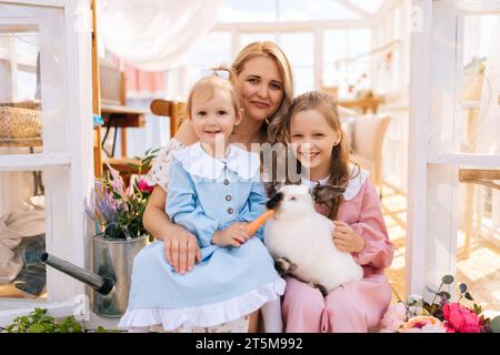 Ritratto della madre sorridente e di due adorabili figlie in abito che abbraccia il coniglio bianco nella casa del gazebo estivo nelle giornate di sole, guardando la macchina fotografica. Foto Stock