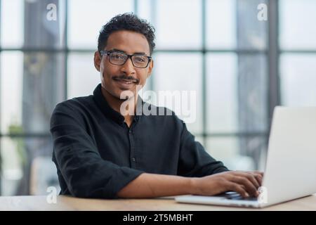 Un uomo d'affari concentrato lavora alla scrivania dell'ufficio aziendale, usando un computer portatile davanti alla finestra. Foto Stock