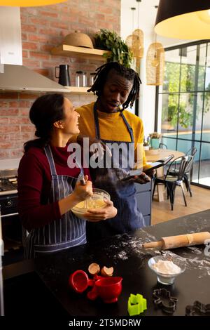 Allegri coppie che preparano biscotti di natale e usano un tablet in una cucina soleggiata Foto Stock