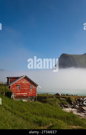 Casa norvegese rossa in un fiordo in una luminosa mattinata di sole, con nuvole di nebbia che si infrangono dal mare. Cielo blu profondo sopra. Foto Stock