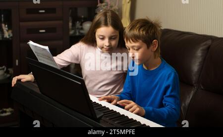 Un ragazzo e una ragazza esercitano il pianoforte insieme a casa, concentrati sullo spartito musicale Foto Stock