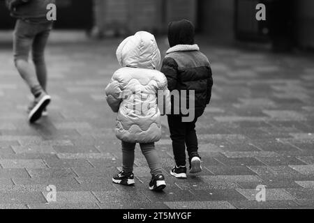 Due bambini piccoli (un ragazzo e una ragazza) vestiti per l'inverno, camminano mano nella mano dietro un adulto che si prende cura di lui Foto Stock